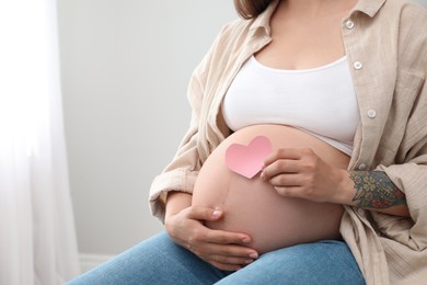 Photo of Pregnant woman with pink paper heart at home, closeup. Space for text