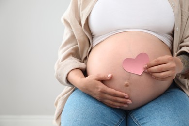 Photo of Pregnant woman with pink paper heart on light background, closeup