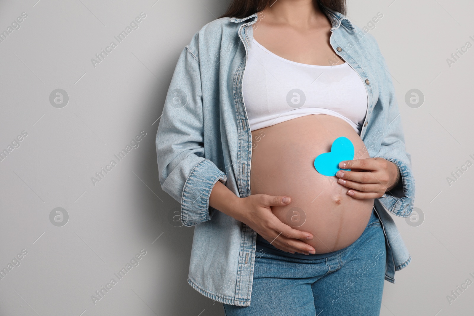 Photo of Pregnant woman with light blue paper heart on gray background, closeup. Space for text