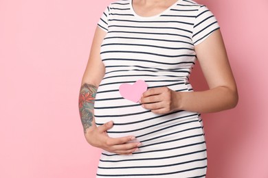 Photo of Pregnant woman with paper heart on pink background, closeup