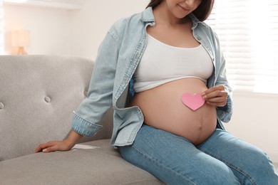 Pregnant woman with pink paper heart on sofa at home, closeup