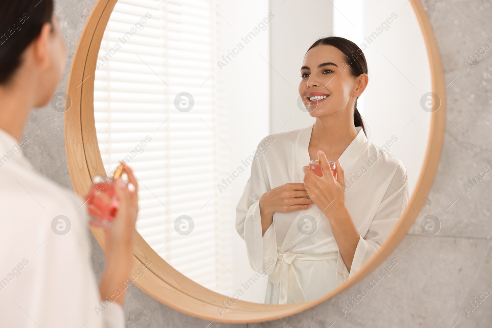 Photo of Smiling woman with bottle of perfume near mirror in bathroom