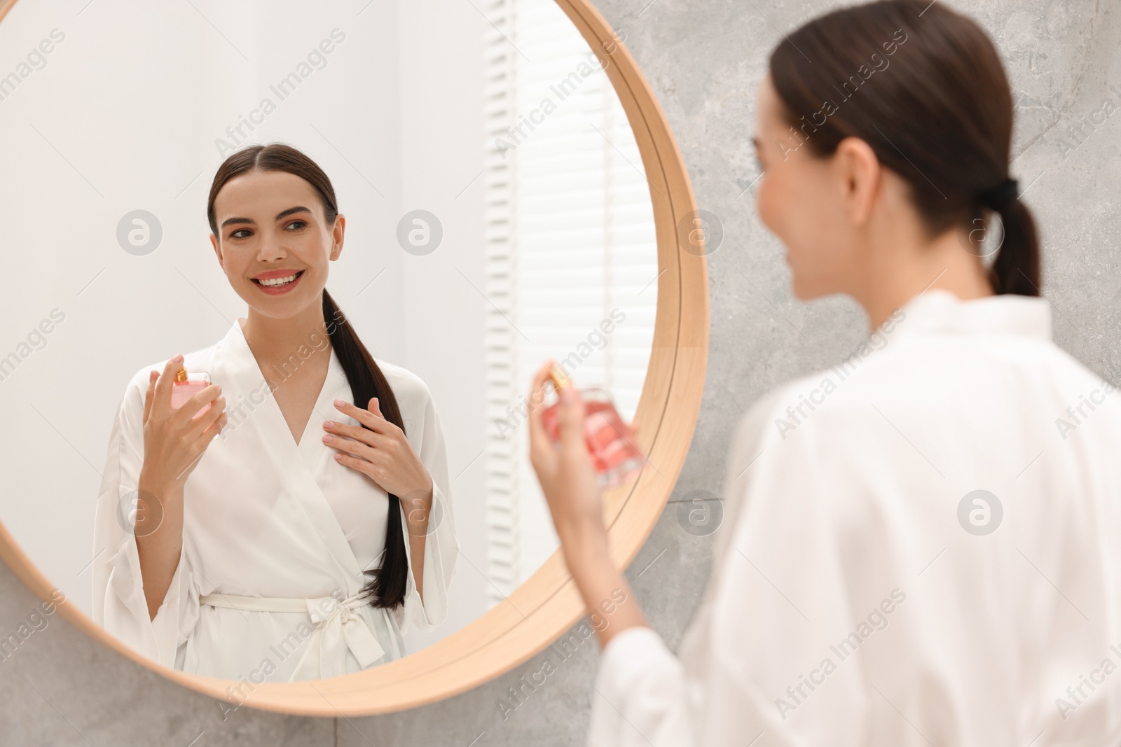 Photo of Smiling woman with bottle of perfume near mirror in bathroom