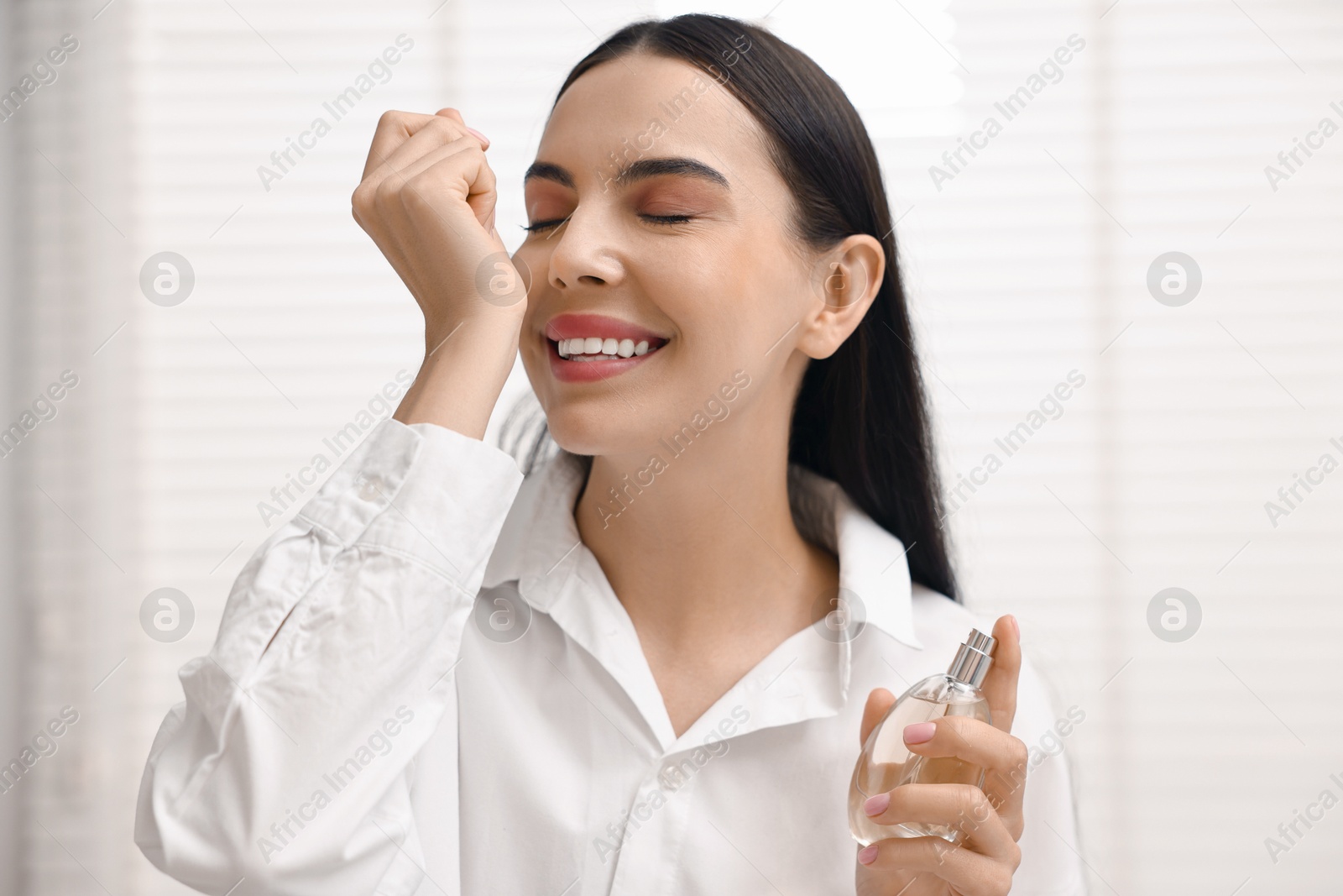 Photo of Smiling woman smelling perfume on wrist at home