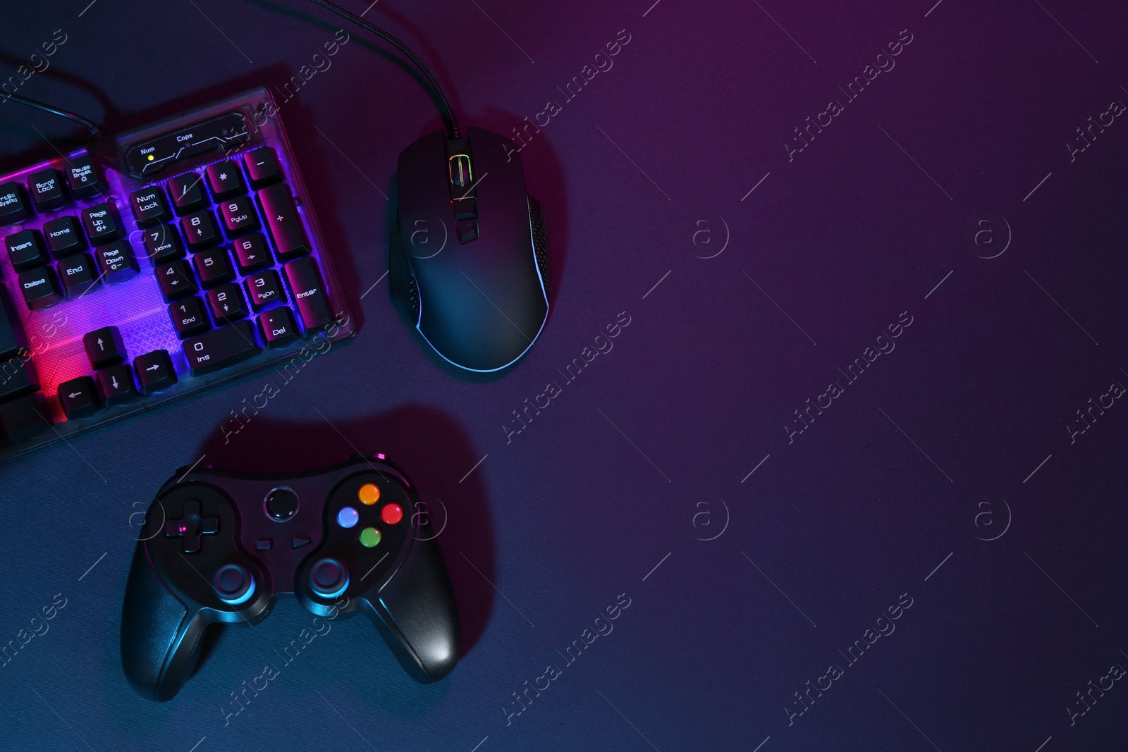 Photo of Computer mouse, RGB keyboard and game controller in neon lights on dark background, flat lay. Space for text