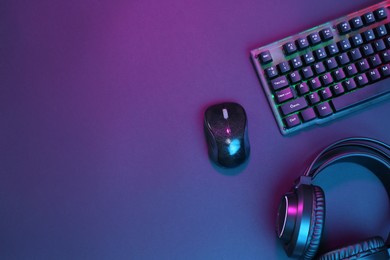 Photo of Computer mouse, RGB keyboard and headset in neon lights on dark background, flat lay. Space for text