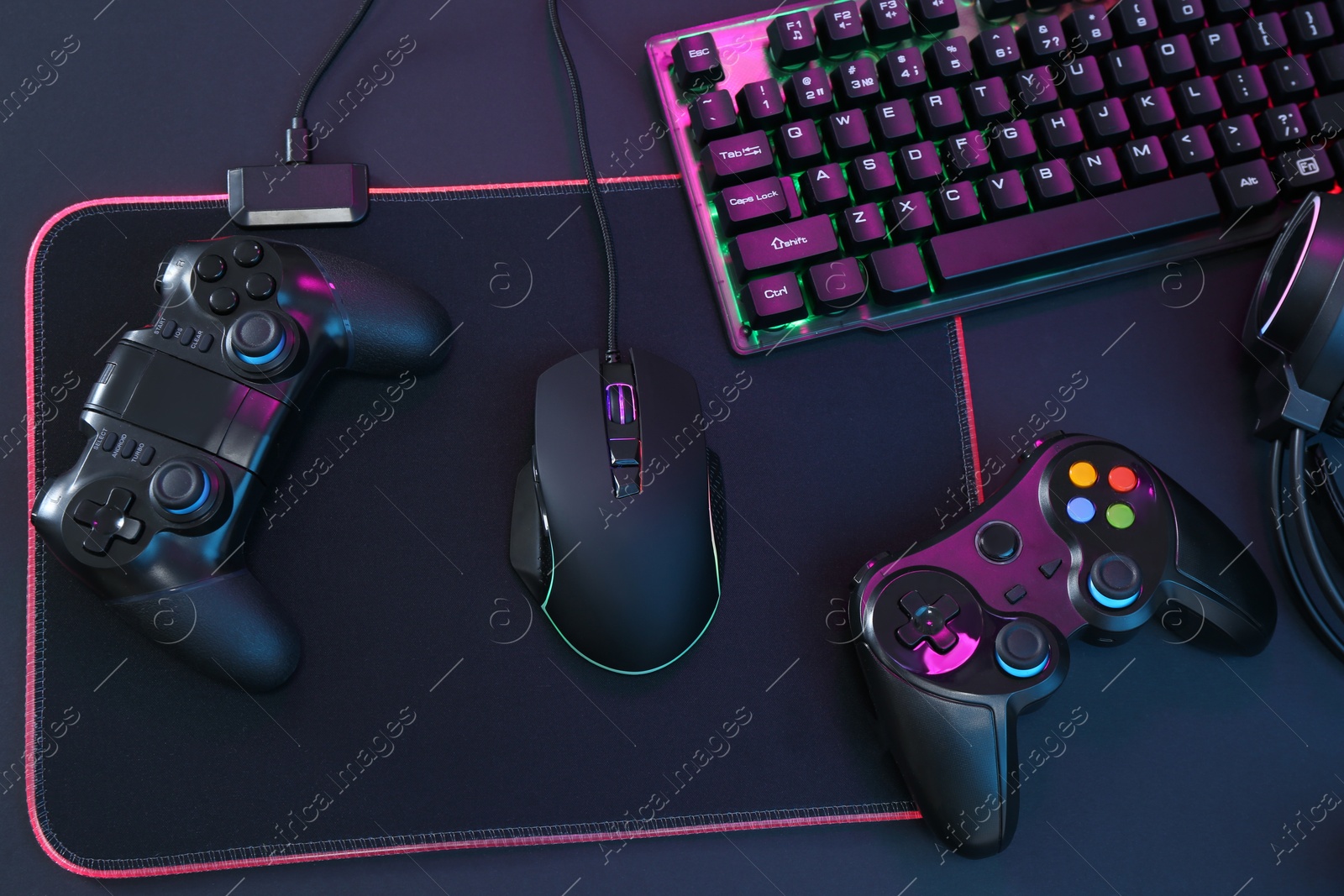 Photo of Computer mouse, RGB keyboard and game controllers in neon lights on dark background, top view