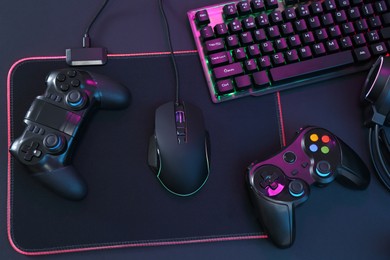 Photo of Computer mouse, RGB keyboard and game controllers in neon lights on dark background, top view