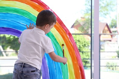Photo of Little boy drawing rainbow on window indoors