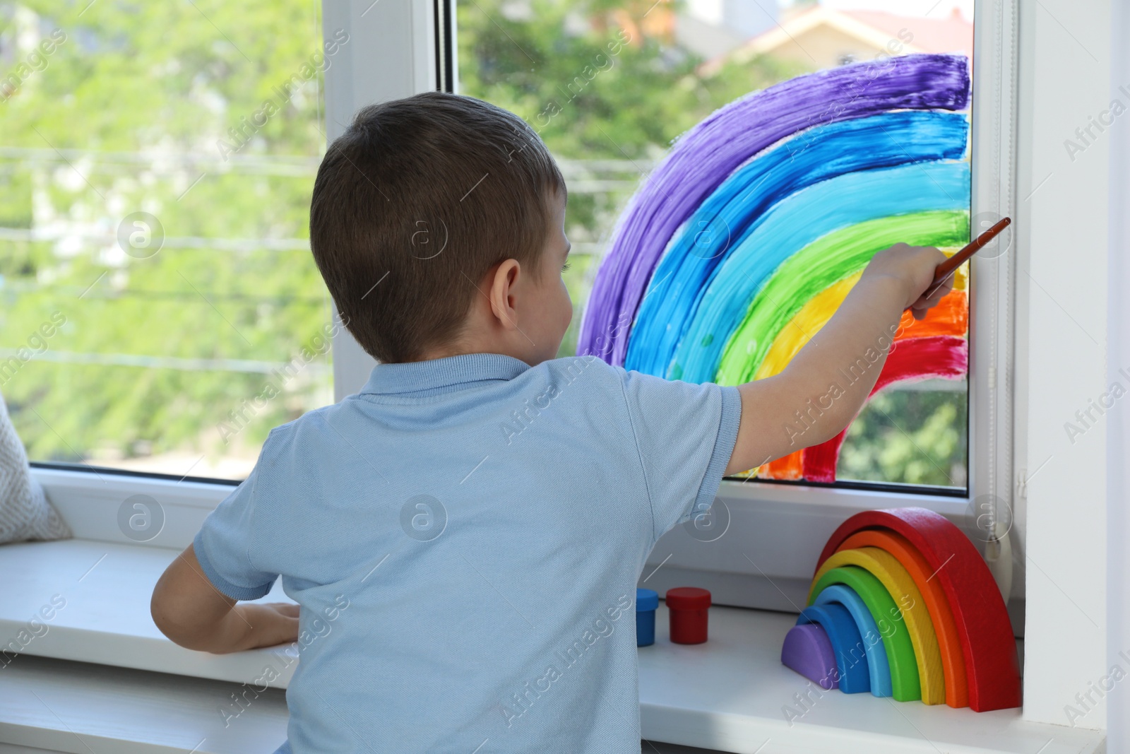 Photo of Little boy drawing rainbow on window indoors