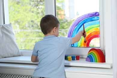 Photo of Little boy drawing rainbow on window indoors