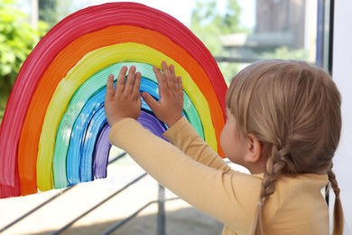 Little girl touching picture of rainbow on window indoors