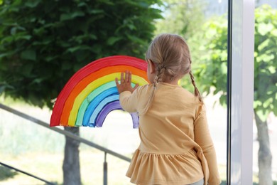 Little girl touching picture of rainbow on window indoors, back view