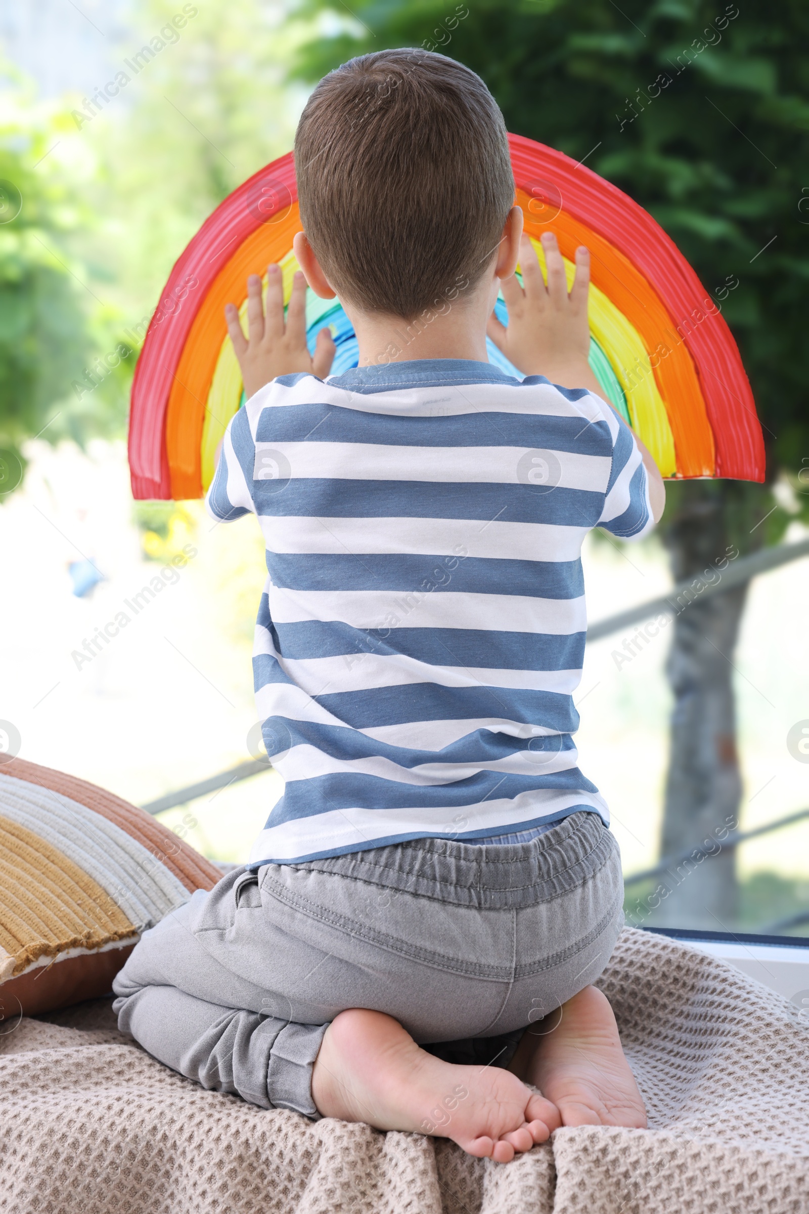 Photo of Little boy touching picture of rainbow on window indoors, back view