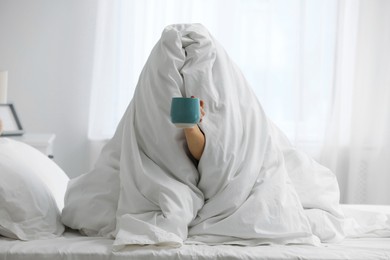 Photo of Woman wrapping in blanket sitting with cup of coffee on bed. Good morning