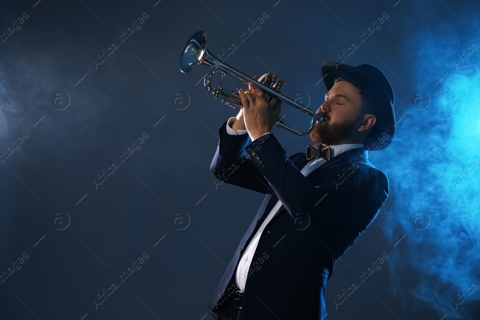 Photo of Professional musician playing trumpet on dark background in blue lights and smoke. Space for text