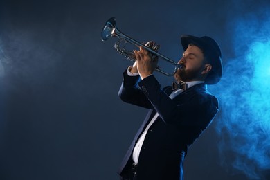 Photo of Professional musician playing trumpet on dark background in blue lights and smoke. Space for text