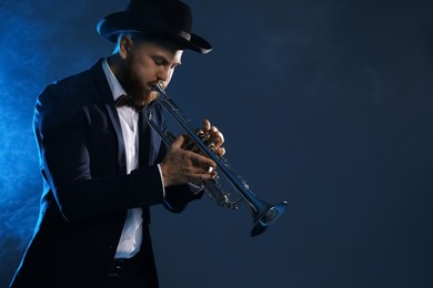 Photo of Professional musician playing trumpet on dark background in blue lights and smoke. Space for text