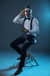 Handsome musician with trumpet sitting on stool against dark background