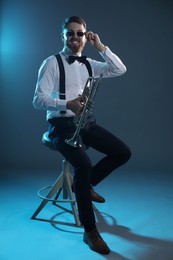 Smiling musician with trumpet sitting on stool against dark background