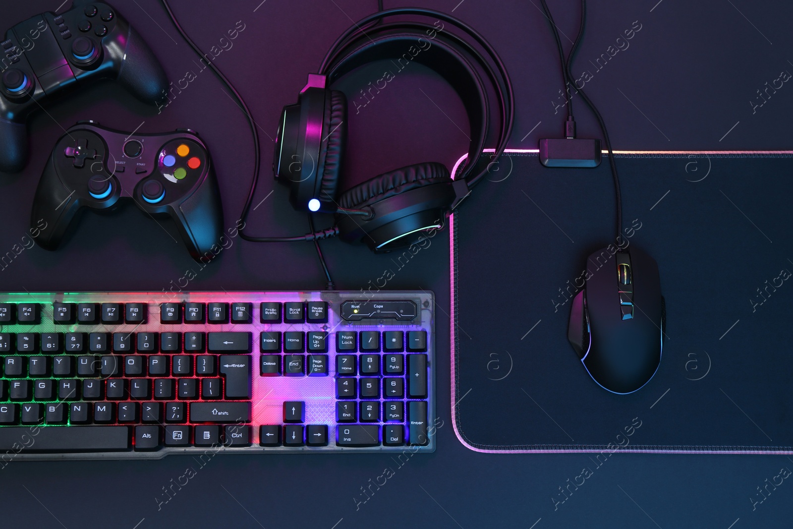 Photo of Computer mouse, RGB keyboard, headset and game controllers in neon lights on dark background, top view