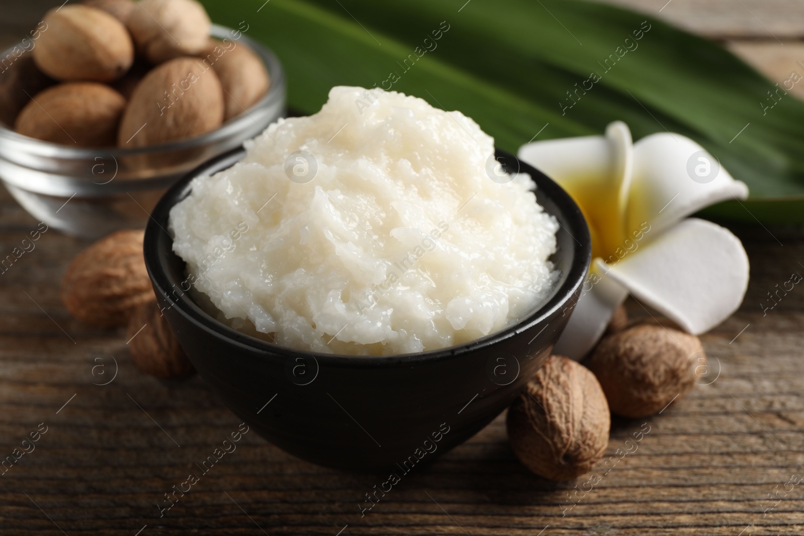 Photo of Natural shea butter in bowl, nuts and plumeria flower on wooden table, closeup