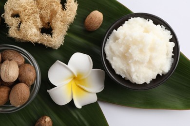 Photo of Natural shea butter in bowl, nuts, plumeria flower, loofah sponge and green leaves on white background, top view