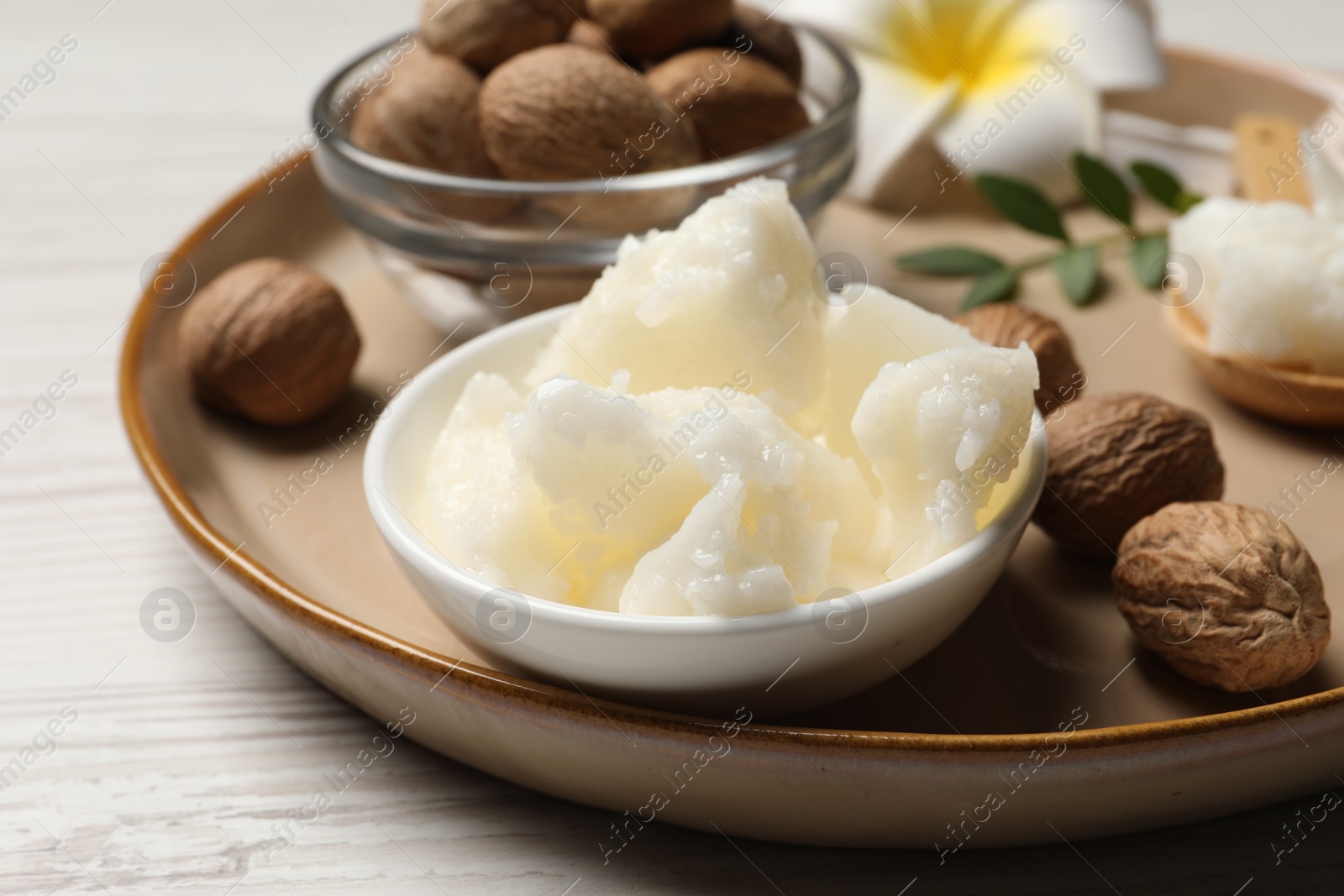 Photo of Natural shea butter in bowl, nuts, plumeria flower and green twig on wooden table, closeup