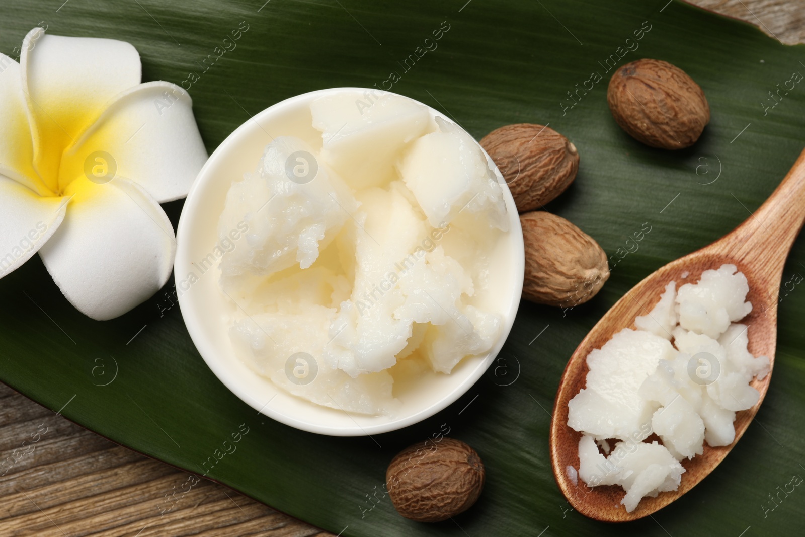 Photo of Natural shea butter in bowl, spoon, nuts, plumeria flower and green leaf on wooden table, top view