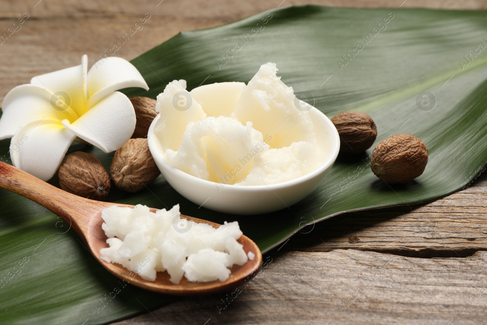 Photo of Natural shea butter in bowl, spoon, nuts, plumeria flower and green leaf on wooden table