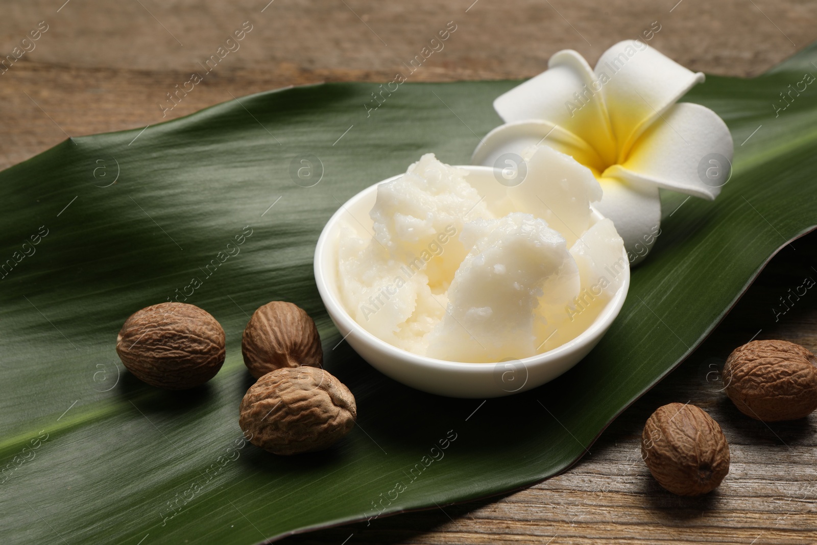 Photo of Natural shea butter in bowl, nuts, plumeria flower and green leaf on wooden table