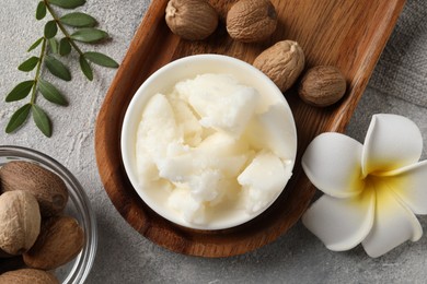 Photo of Natural shea butter in bowl, nuts, green leaves and plumeria flower on grey table, top view