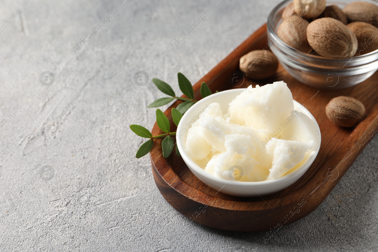 Photo of Natural shea butter in bowl, nuts, green leaves and plumeria flower on grey table, space for text