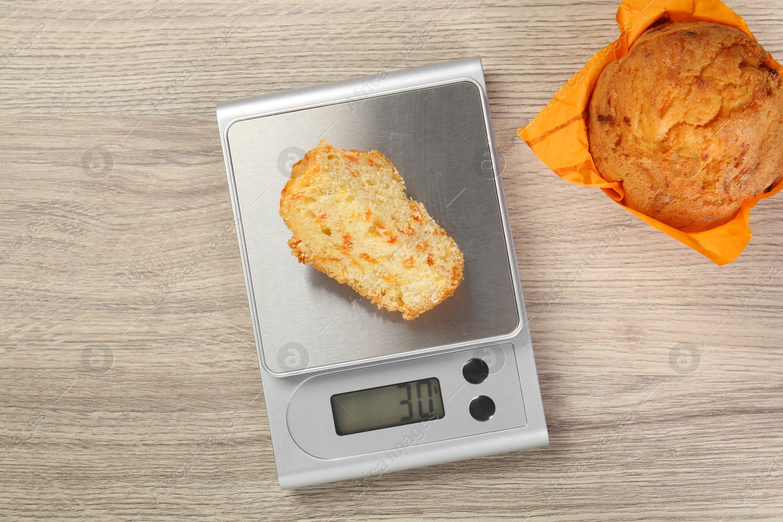 Photo of Kitchen scale with piece of muffin on light wooden table, flat lay