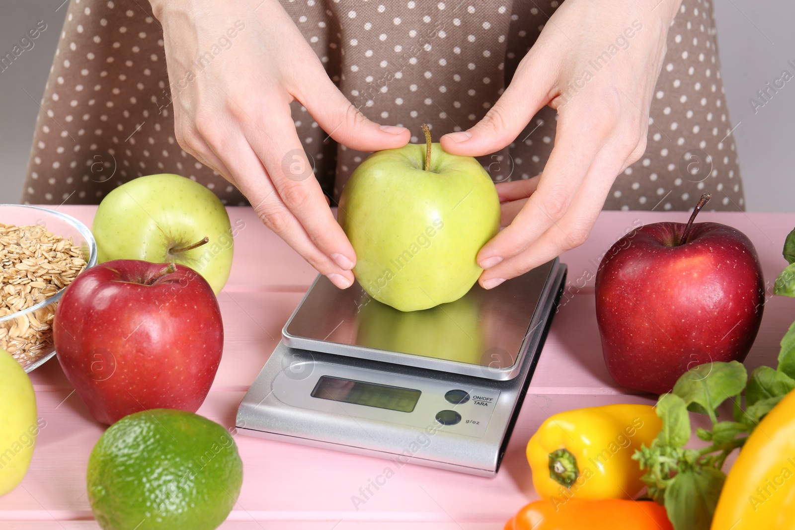 Photo of Woman weighing apple on kitchen scale at pink wooden table, closeup