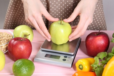 Photo of Woman weighing apple on kitchen scale at pink wooden table, closeup