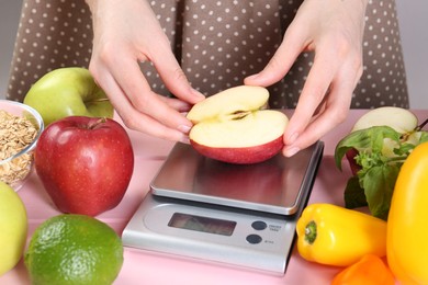 Photo of Woman weighing half of apple on kitchen scale at pink wooden table, closeup
