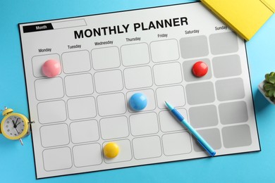 Photo of Timetable. Monthly planner, pen, drawing pins and alarm clock on light blue background, top view