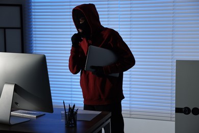 Thief wearing balaclava stealing laptop in office at night. Burglary