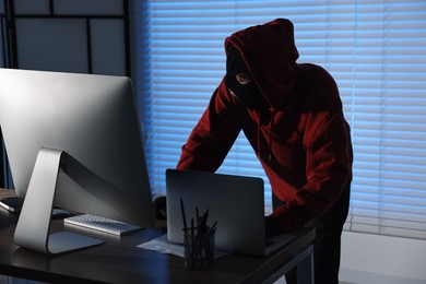 Thief wearing balaclava stealing laptop in office at night. Burglary