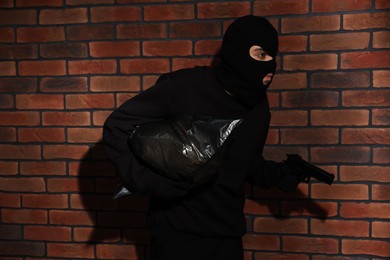 Thief in balaclava with gun and bag against red brick wall