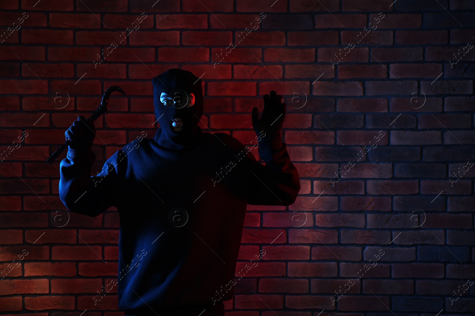 Photo of Thief in balaclava raising hands with crowbar in color lights against red brick wall. Space for text