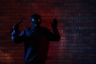 Thief in balaclava raising hands with crowbar in color lights against red brick wall. Space for text