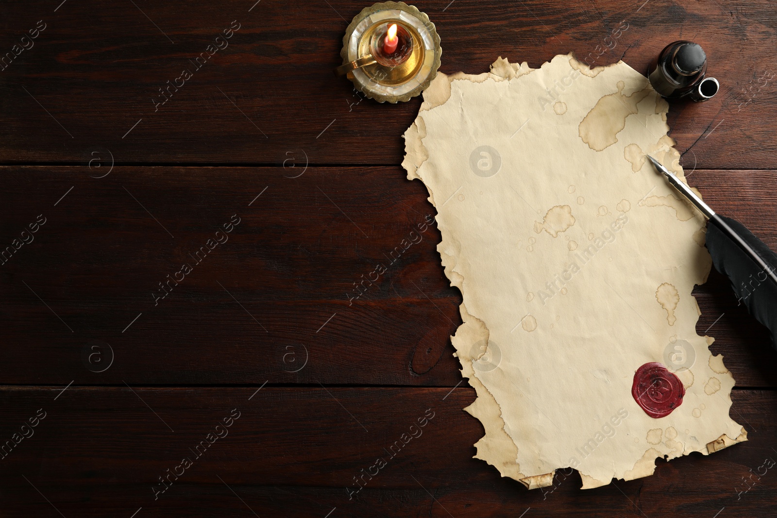 Photo of Sheet of old parchment paper with wax stamp, black feather, inkwell and candle on wooden table, flat lay. Space for text