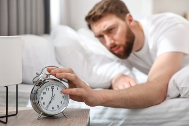 Sleepy young man turning off alarm clock in bedroom at morning, selective focus