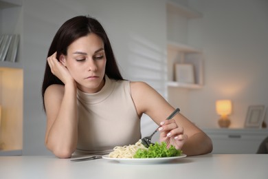 Eating disorder. Sad woman holding fork near plate with spaghetti at white table indoors