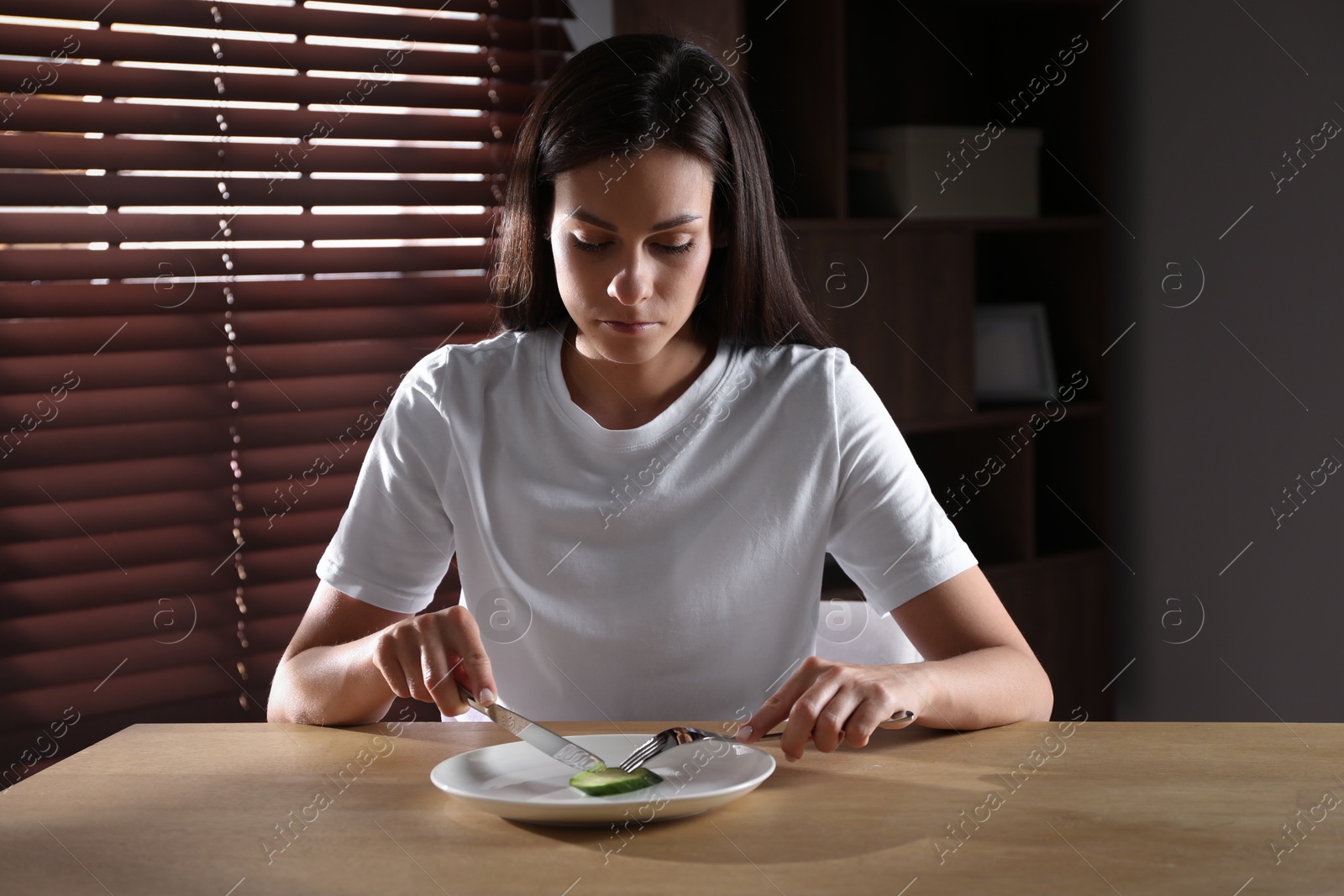 Photo of Eating disorder. Sad woman cutting cucumber at wooden table indoors