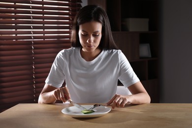 Eating disorder. Sad woman cutting cucumber at wooden table indoors