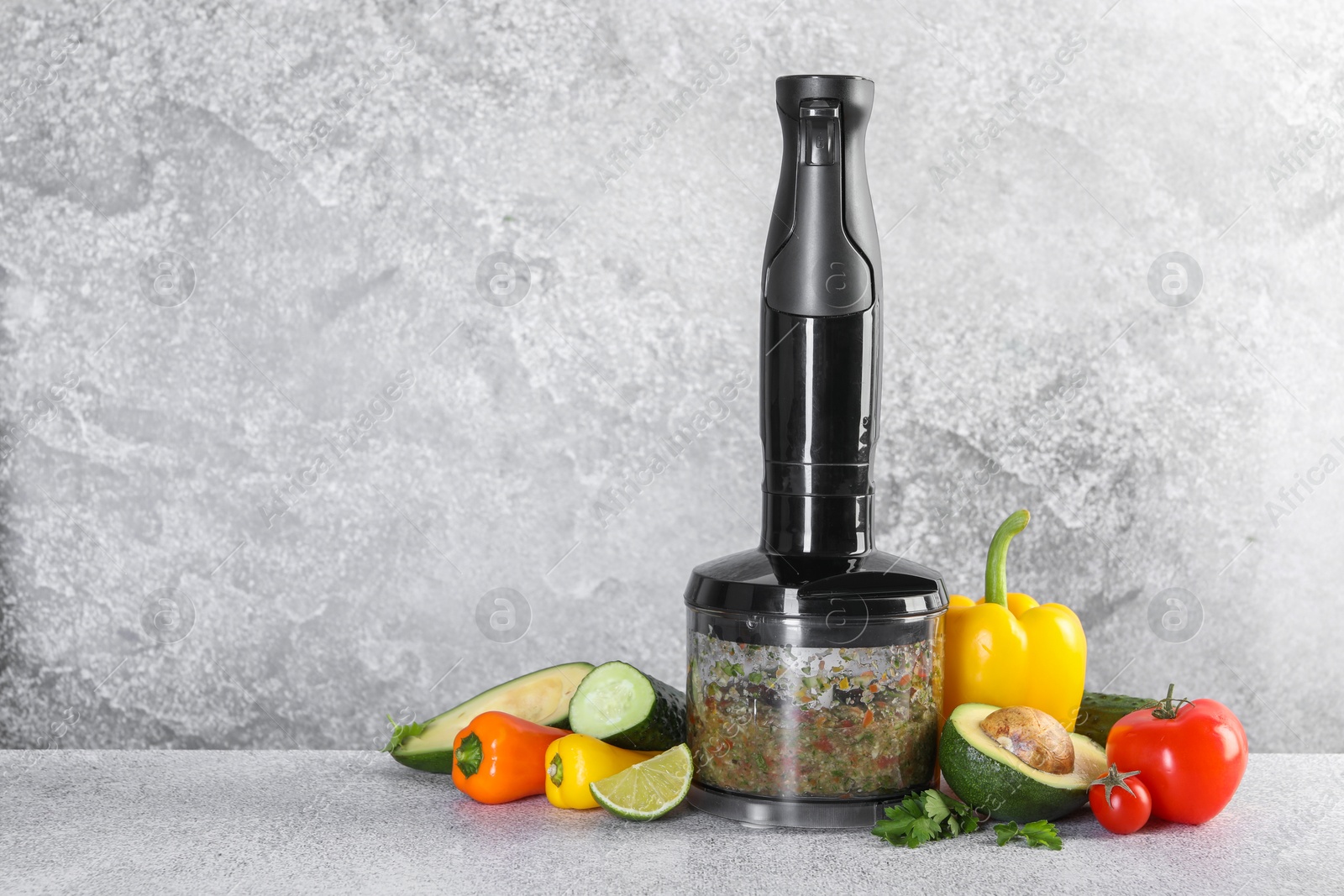 Photo of Hand blender with mixture of ingredients and fresh products on light grey table, space for text