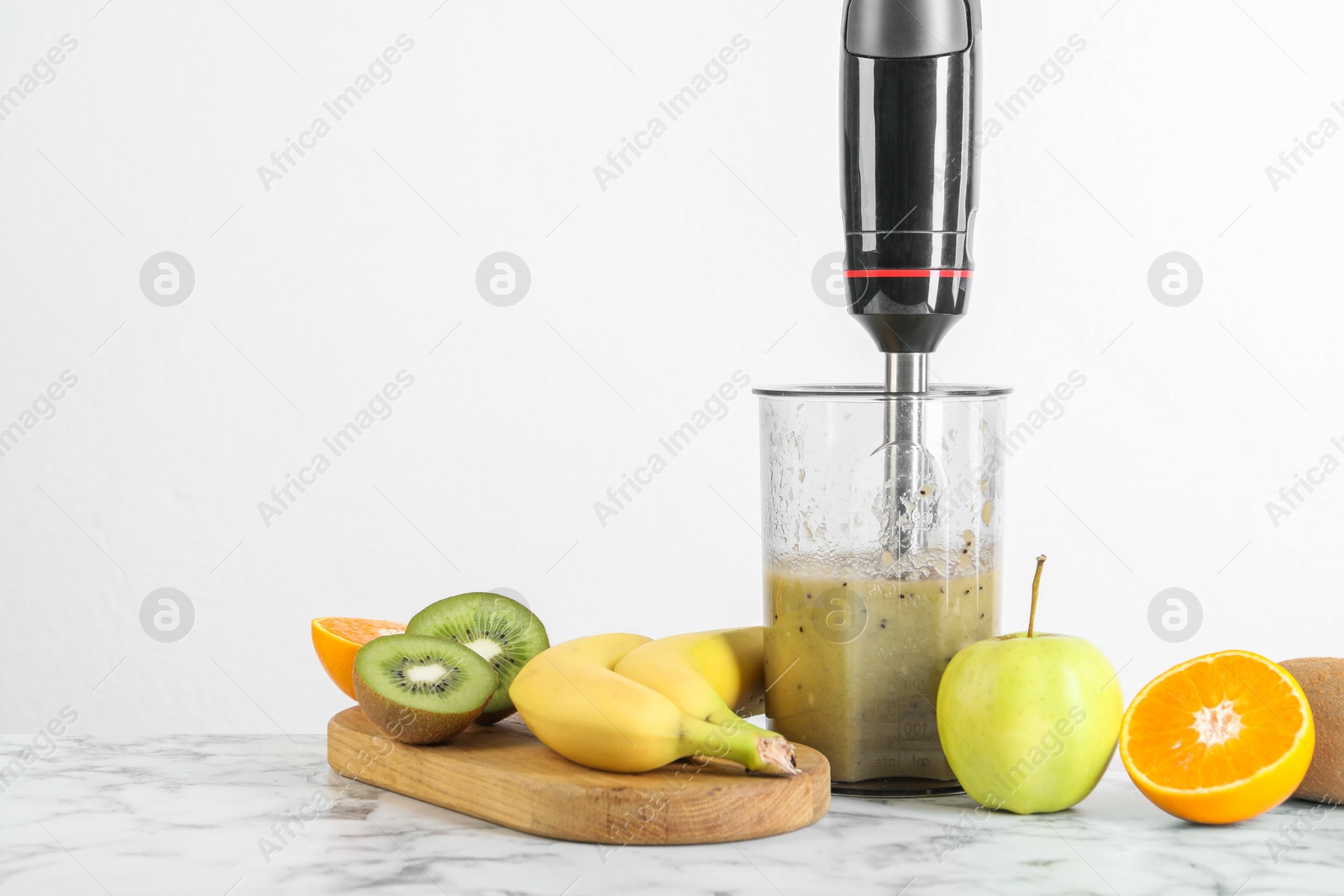 Photo of Hand blender with mixture of ingredients and fresh fruits on white marble table